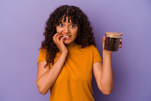 Mujer joven de raza mixta sosteniendo una botella de granos de café aislada en la pared púrpura mordiéndose las uñas, nerviosa y muy ansiosa.