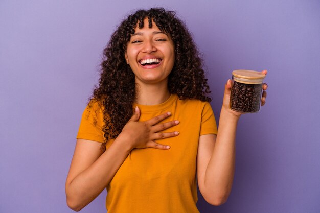 Mujer joven de raza mixta sosteniendo una botella de granos de café aislada en la pared púrpura se ríe a carcajadas manteniendo la mano en el pecho.
