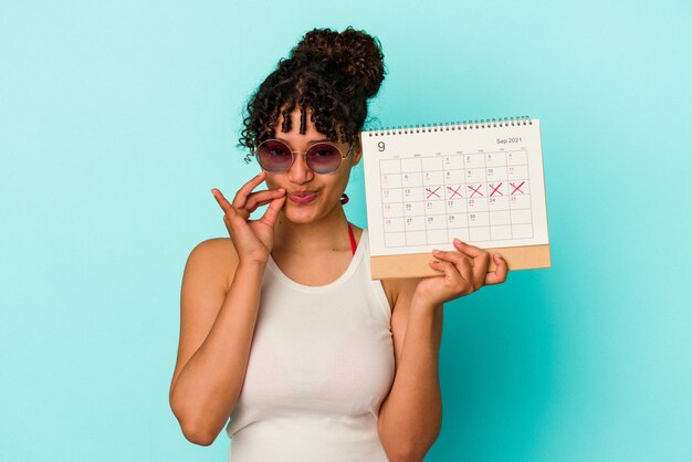 Mujer joven de raza mixta sosteniendo el calendario aislado en la pared azul con los dedos en los labios manteniendo un secreto.