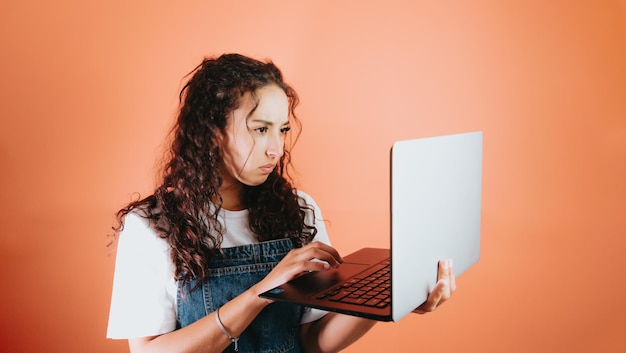 Mujer joven de raza mixta sosteniendo una computadora portátil y preocupada por algo que está viendo en la pantalla, fondo naranja fondo extraíble, imagen mínima, espacio de copia, situaciones básicas