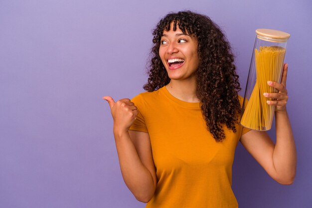 Mujer joven de raza mixta sosteniendo espaguetis aislados en puntos de pared púrpura con el dedo pulgar lejos, riendo y despreocupado.