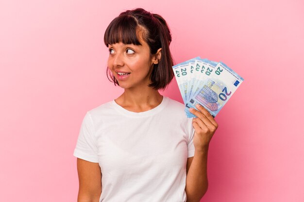 Mujer joven de raza mixta sosteniendo facturas aisladas sobre fondo rosa se ve a un lado sonriente, alegre y agradable.