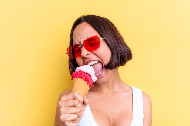 Mujer joven de raza mixta sosteniendo un helado aislado en la pared amarilla