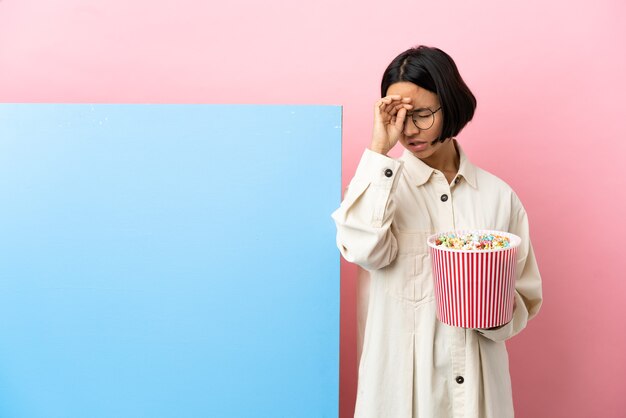 Mujer joven de raza mixta sosteniendo palomitas de maíz con una gran pancarta sobre una pared aislada con expresión cansada y enferma