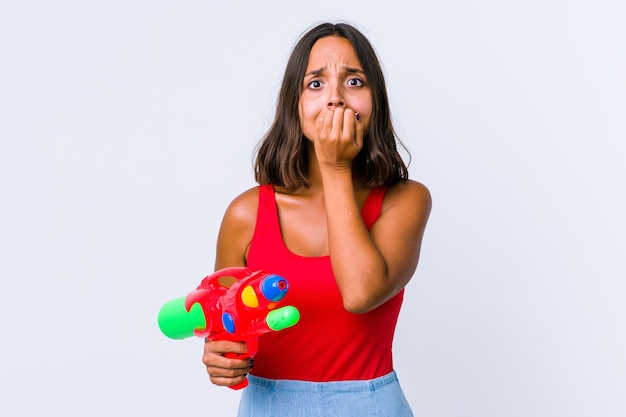 Mujer joven de raza mixta sosteniendo una pistola de agua aislada mordiendo las uñas, nerviosa y muy ansiosa.