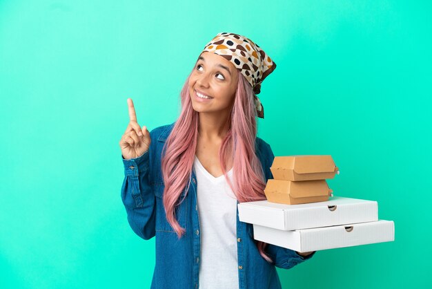 Mujer joven de raza mixta sosteniendo pizzas y hamburguesas aisladas sobre fondo verde apuntando hacia una gran idea