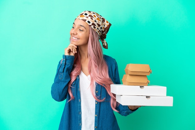 Mujer joven de raza mixta sosteniendo pizzas y hamburguesas aisladas sobre fondo verde mirando hacia el lado y sonriendo