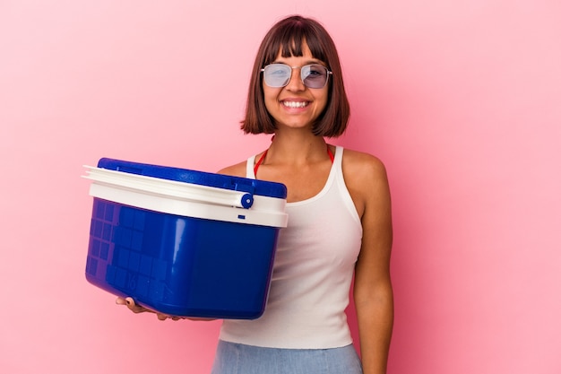 Mujer joven de raza mixta sosteniendo un refrigerador aislado sobre fondo rosa feliz, sonriente y alegre.