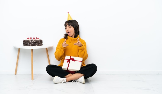 Mujer joven de raza mixta sosteniendo un regalo sentado en el suelo aislado sobre fondo blanco apuntando al frente y sonriendo