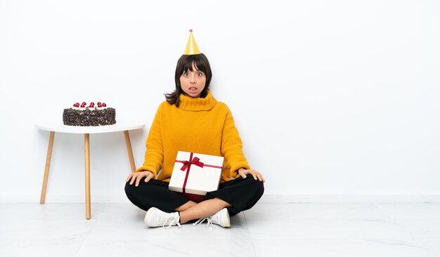 Mujer joven de raza mixta sosteniendo un regalo sentado en el suelo aislado sobre fondo blanco con expresión facial sorpresa