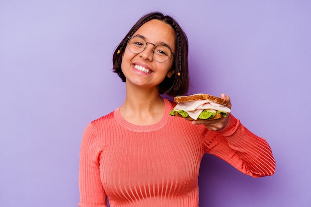 Mujer joven de raza mixta sosteniendo un sándwich aislado sobre fondo púrpura feliz, sonriente y alegre.