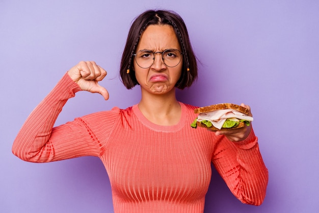 Mujer joven de raza mixta sosteniendo un sándwich aislado sobre fondo púrpura mostrando un gesto de aversión, pulgares hacia abajo. Concepto de desacuerdo.