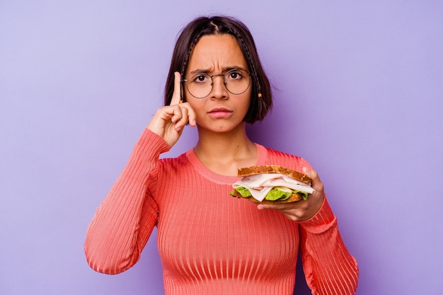 Mujer joven de raza mixta sosteniendo un sándwich aislado sobre fondo púrpura señalando la sien con el dedo, pensando, centrado en una tarea.