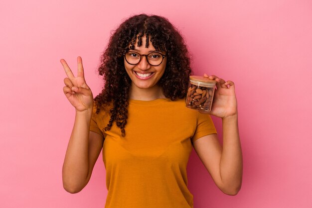 Mujer joven de raza mixta sosteniendo un tarro de almendras aislado sobre fondo rosa mostrando el número dos con los dedos.