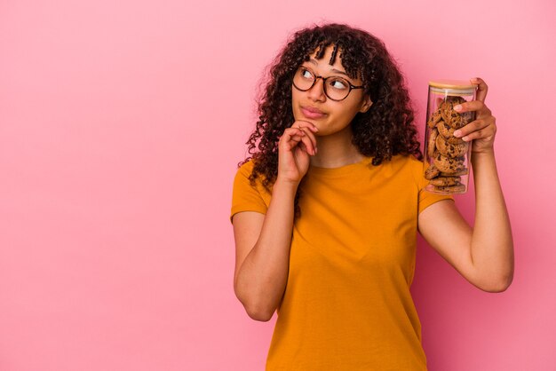 Mujer joven de raza mixta sosteniendo un tarro de galletas aislado sobre fondo rosa mirando hacia los lados con expresión dudosa y escéptica.