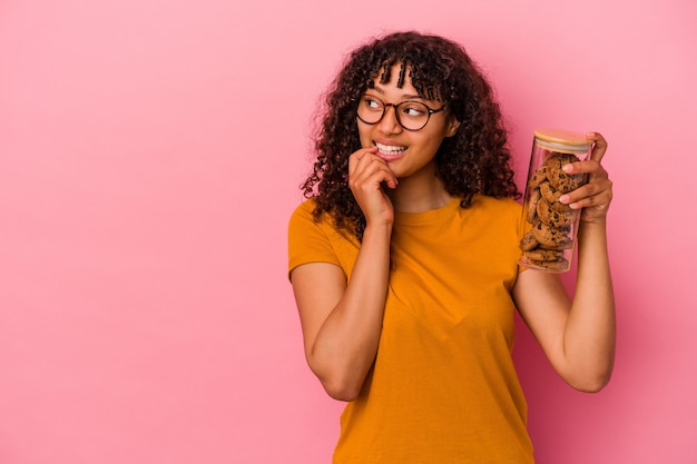 Mujer joven de raza mixta sosteniendo un tarro de galletas aislado sobre fondo rosa relajado pensando en algo mirando un espacio de copia.