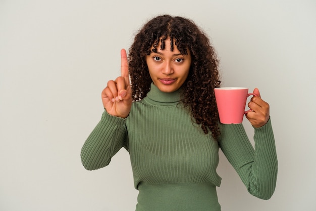 Mujer joven de raza mixta sosteniendo una taza aislada sobre fondo blanco mostrando el número uno con el dedo.