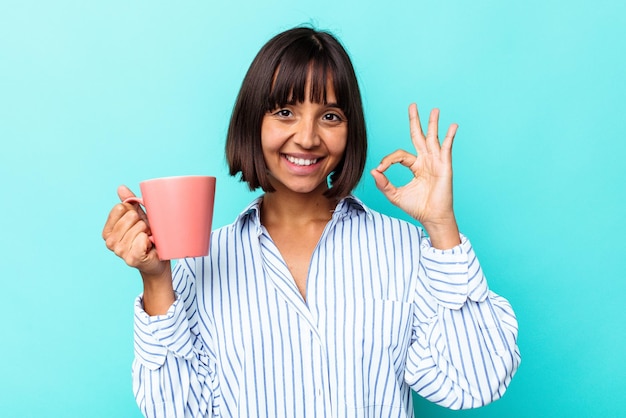 Mujer joven de raza mixta sosteniendo una taza rosa aislada sobre fondo azul alegre y confiado mostrando gesto ok.