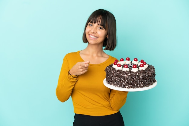 Mujer joven de raza mixta sosteniendo la torta de cumpleaños apuntando hacia el frente con expresión feliz