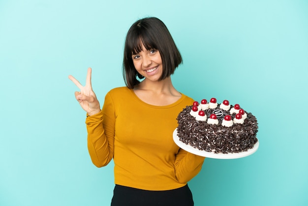 Mujer joven de raza mixta sosteniendo la torta de cumpleaños sonriendo y mostrando el signo de la victoria