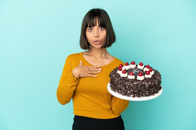 Mujer joven de raza mixta sosteniendo la torta de cumpleaños sorprendida y conmocionada mientras mira a la derecha