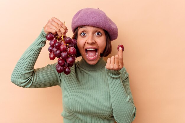 Mujer joven de raza mixta sosteniendo uvas aisladas sobre pared beige