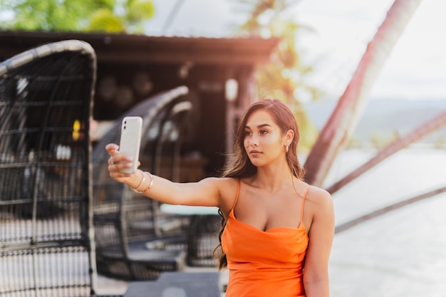 Mujer joven de raza mixta tomando un selfie en un teléfono celular en vestido naranja de verano