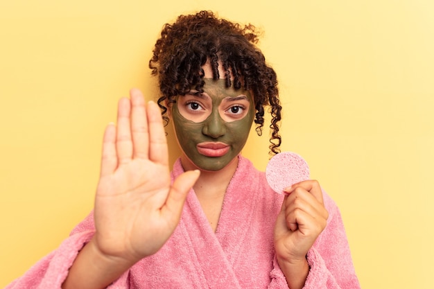 Mujer joven de raza mixta vistiendo una bata de baño sosteniendo una esponja desmaquillante aislada sobre fondo amarillo de pie con la mano extendida mostrando la señal de stop, impidiéndote.