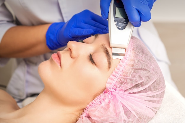 Mujer joven recibiendo limpieza de la piel facial por equipos ultrasónicos en el salón médico