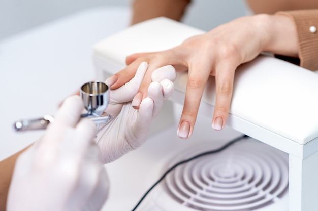 Mujer joven recibiendo manicura con aerógrafo en salón de uñas.  procedimiento para rociar pintura en las uñas.