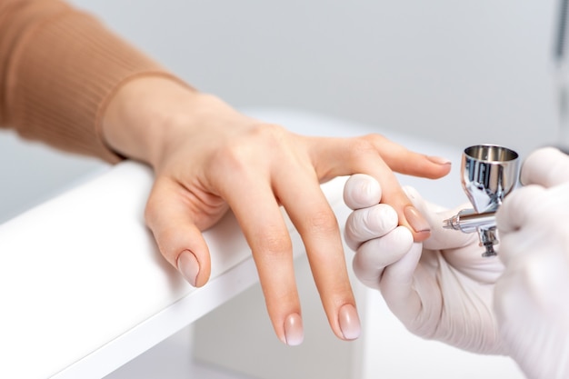 Mujer joven recibiendo manicura con aerógrafo en salón de uñas. Procedimiento para rociar pintura en las uñas.