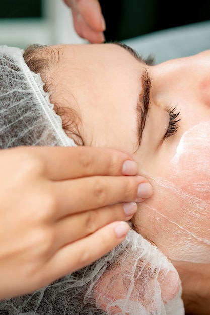 Mujer joven recibiendo masaje facial con los ojos cerrados por esteticista en salón de belleza