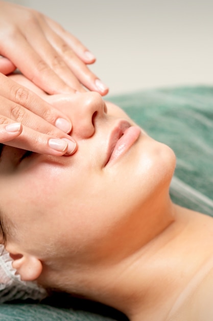 Mujer joven recibiendo masaje facial con los ojos cerrados por esteticista en salón de belleza