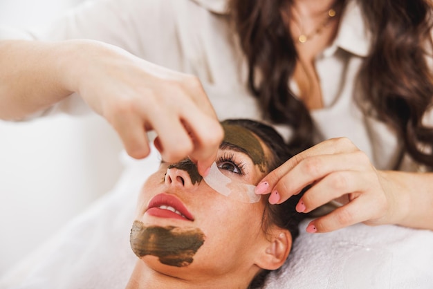 Mujer joven recibiendo parches en los ojos y tratamiento facial con barro de algas en el spa.