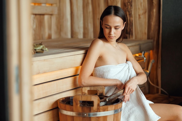 Mujer joven recoge agua de un balde de madera en una sauna