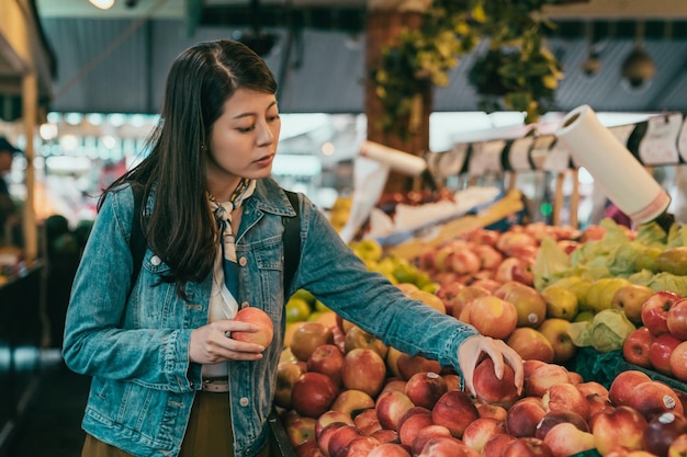 mujer joven recogiendo manzanas en el mercado de agricultores original, está comparando dos manzanas y va a volver a poner una en el estante