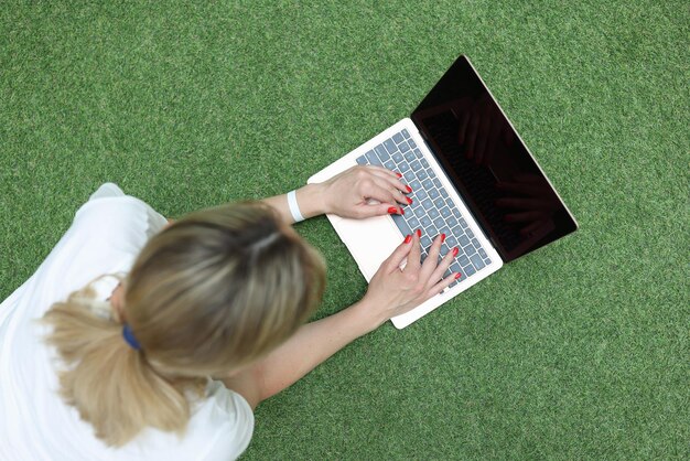 Mujer joven recostada sobre la hierba verde y escribiendo en la vista superior del teclado de la computadora portátil