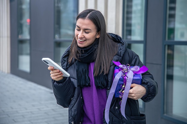 Mujer joven con un regalo morado en las manos usa un teléfono inteligente afuera