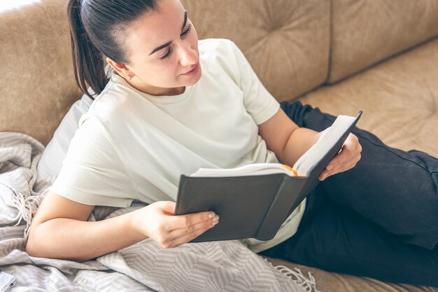 Una mujer joven se relaja en casa tendida en el sofá con un libro en las manos