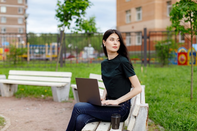 Mujer joven relajada en una camiseta verde y con una mochila sentado en un banco de madera tomando café y navegando en su computadora portátil