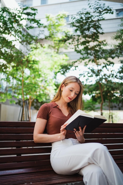 Mujer joven relajada leyendo un libro mientras se sienta en un banco en el campus universitario