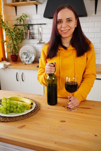 Mujer joven relajada posa con una copa de vino tinto de pie en la mesa de la cocina Hembra adulta descansando con alcohol y frutas en la cocina