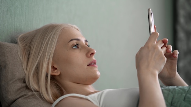 Mujer joven relajada que usa teléfonos inteligentes navegando en las redes sociales, revisando noticias, jugando juegos móviles o enviando mensajes de texto sentados en el sofá.