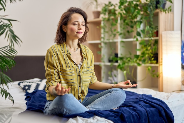 Mujer joven relajada sentada en la cama en posición de loto meditando en casa