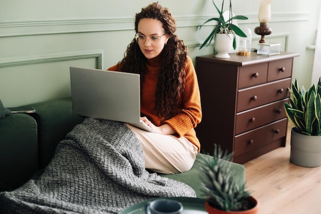 Mujer joven relajada sentada en un sofá sosteniendo un teléfono móvil usando tecnología de teléfono celular y computadora portátil haciendo