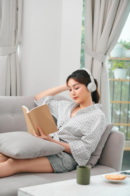 Mujer joven relajada en el sofá con auriculares leyendo un libro