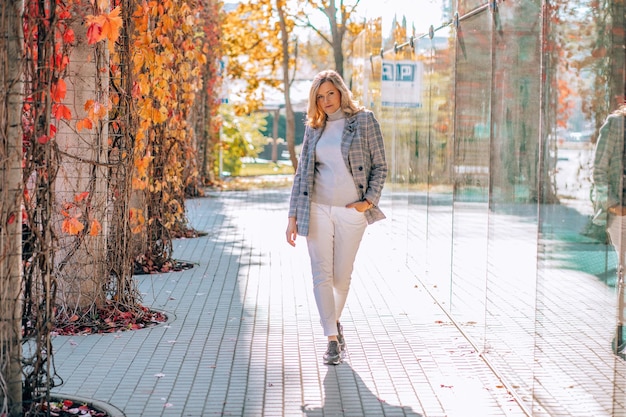 Mujer joven relajada con suéter ligero, jeans blancos y chaqueta gris caminando al aire libre posiblemente en la ciudad o pueblo en un cálido día soleado a principios de otoño con su naturaleza colorida y hojas rojas y naranjas
