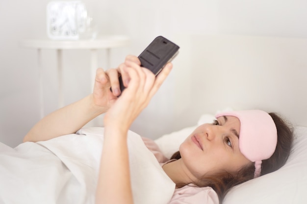 Mujer joven relajada con un teléfono inteligente por la mañana en la cama en casa. Niña feliz y sonriente con un teléfono móvil. Mujer enviando mensajes de texto, revisando aplicaciones sociales, haciendo videollamadas o jugando con el teléfono inteligente.