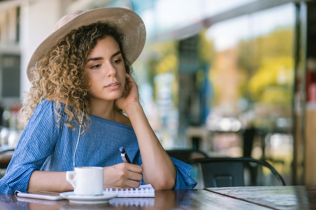 Mujer joven relajándose y escribiendo en un cuaderno mientras bebe una taza de café en una cafetería.