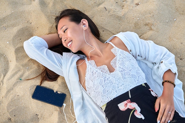 Mujer joven relajante en auriculares con teléfono inteligente en la vista superior de la arena